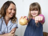 A smiling woman and her young daughter. The daughter is holding a potato in one hand and her fist outstretched, to compare the size of the potato and her hand.