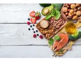 A selection of foods that contain healthy fats on a white table, including avocado, salmon, nuts and oil