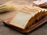 Four slices of white bread on a wooden chopping board, with strands of wheat in the background.