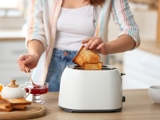 A woman taking a piece of toast out of a white toaster. She is also spooning some jam out of a jar to spread on the toast.